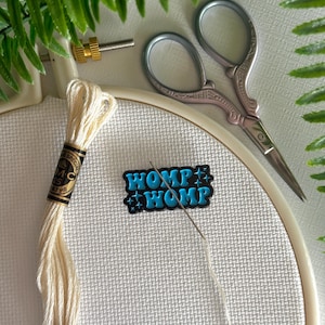 May include: A close-up of a white cross-stitch fabric stretched in a beige embroidery hoop. The image includes a spool of cream-colored thread, a needle, small silver scissors, and a pin that says "WOMP WOMP" in blue and white.
