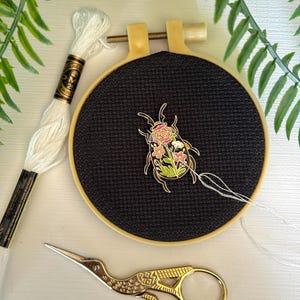 May include: A black cross-stitch embroidery hoop with a floral beetle design. White embroidery floss and gold scissors are next to the hoop. The beetle is decorated with pink, yellow, and green flowers. The embroidery hoop is made of wood.