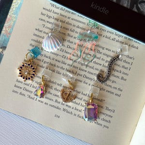 May include: A collection of decorative Kindle page holders featuring various charms. The charms include a seashell, a heart, a crystal, a nautical wheel, and an octopus. The page holders are clear and attached to a book.