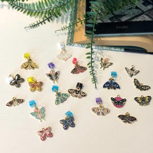 May include: A collection of colorful butterfly charms with various designs and colors, including gold, pink, blue, and green. Each charm has a small clasp. The word "kindle" is visible on a black e-reader.