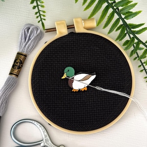May include: A cross-stitch embroidery hoop with a black fabric background features a colorful duck design. Gray embroidery floss and small silver scissors are next to the hoop. Green fern leaves are in the background.