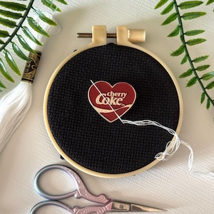 May include: A black cross-stitch project in a beige embroidery hoop, featuring a red heart-shaped Cherry Coke pin. White thread and a pair of pink embroidery scissors are also present. Green fern-like leaves are in the background.