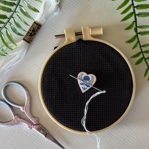 May include: A black cross-stitch hoop with a heart-shaped needle minder featuring a blue floral design. White embroidery floss, a pair of pink scissors, and a spool of white thread are also visible, suggesting a crafting theme.