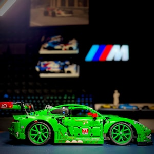 May include: A bright green toy race car constructed from interlocking plastic building blocks. The vehicle features a large rear spoiler, multi-spoke green wheels, and the number 77 printed on the side door panel. It has a low-profile design.