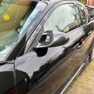 Könnte beinhalten: Ein glänzend schwarzes Sportwagen mit geschwungener Dachlinie und Seitenspiegel. Die Karosserie des Autos spiegelt die Umgebung wider, und die Seitenscheibe ist leicht getönt. Ein gelber Aufkleber ist auf der Windschutzscheibe zu sehen.
