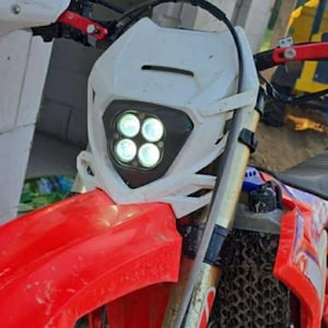 May include: Close-up of a red and white dirt bike. The front of the bike features a white headlight housing with four circular lights. The front fender is red, and the handlebars are visible. The bike appears to be outdoors.