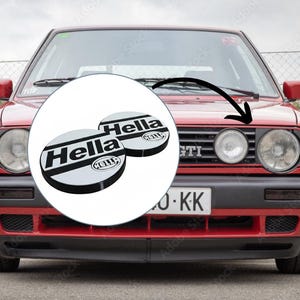 Op de afbeelding: Een rode hatchback met een zwarte grille en ronde koplampen. Een cirkelvormige grafische overlay toont twee witte en zwarte ronde Hella-koplampafdekkingen met het Hella-logo, met een zwarte pijl die naar de grille van de auto wijst.