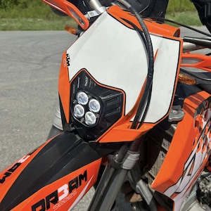 May include: Close-up of an orange and black motorcycle. The motorcycle has a white front panel with the KTM logo and four round headlights. The text "DR3AM Series" is visible on the front.