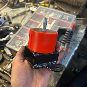 Puede incluir: Un soporte antivibración cilíndrico de goma roja con un perno metálico roscado central que sobresale por la parte superior, unido a un soporte de acero negro con recubrimiento en polvo con base rectangular y un orificio de montaje lateral.
