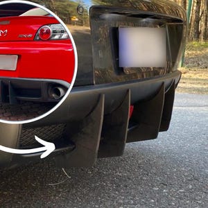 May include: A black car's rear bumper with a diffuser and exhaust is shown. A red sports car is visible in the background. The car is parked on a paved road with trees in the background.