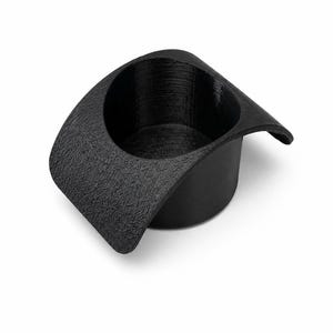 Puede incluir: Un soporte cilíndrico negro para vasos con un acabado texturizado. El objeto presenta un borde ancho y curvado que se extiende hacia afuera en un lado, creando una forma distintiva diseñada para encajar en una abertura circular o superficie empotrada.