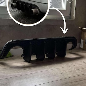 May include: A black car rear diffuser, a vehicle part designed to manage airflow. The diffuser is shown on a wooden floor, with a close-up of the car's exhaust pipes in the background. The diffuser has a curved design.