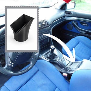 Op de afbeelding: Een zwart, 3D-geprint kunststof auto-interieuraccessoire met een rechthoekige opening aan de bovenzijde die taps toeloopt naar een cilindrische basis. Dit onderdeel is ontworpen voor plaatsing in de middenconsole, zoals aangegeven door de witte pijl.
