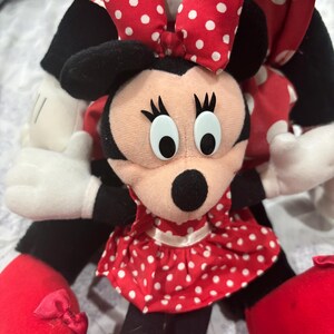May include: A Minnie Mouse plush doll with a red and white polka dot dress and bow. The doll has black ears, a black nose, and large white eyes. The doll's shoes are red with small bows.
