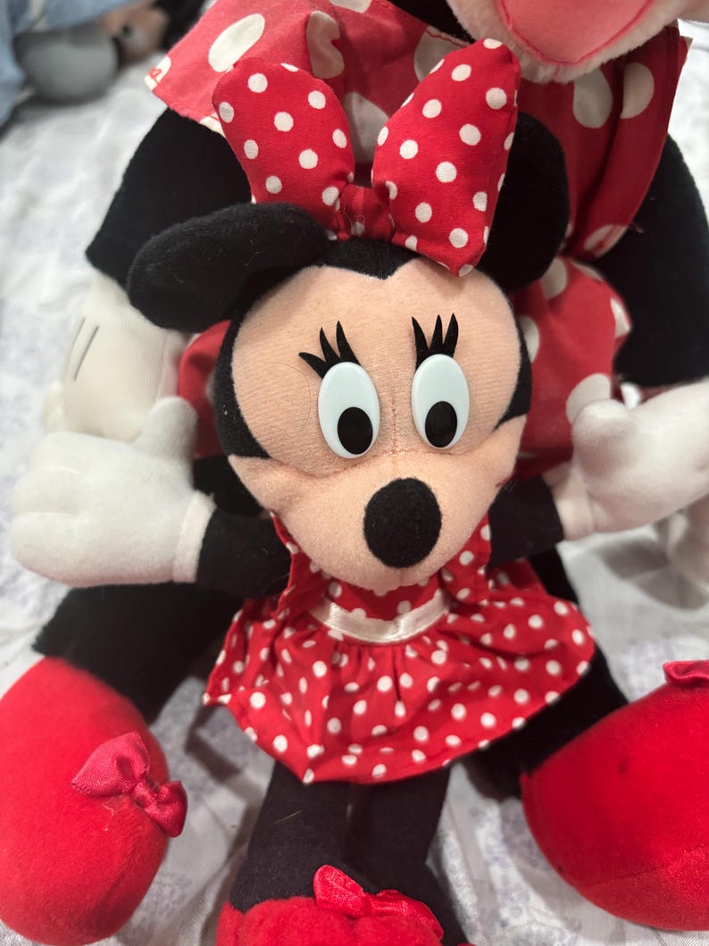 May include: A Minnie Mouse plush doll with a red and white polka dot dress and bow. The doll has black ears, a black nose, and large white eyes. The doll's shoes are red with small bows.