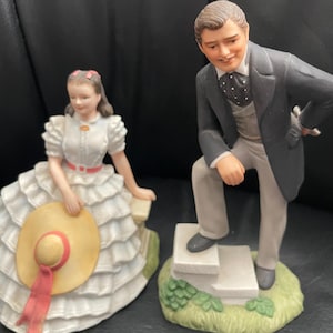 Puede incluir: Dos figuras de porcelana que representan a un hombre y una mujer. La mujer está sentada, con un vestido blanco y un sombrero. El hombre está de pie, con un traje. Ambas figuras están sobre una base con hierba.