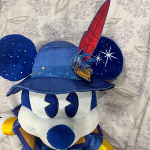 May include: A plush Mickey Mouse dressed as a sorcerer, wearing a blue hat with a red feather and a gold emblem. The ears are dark blue with white star accents. The outfit is blue and gold.