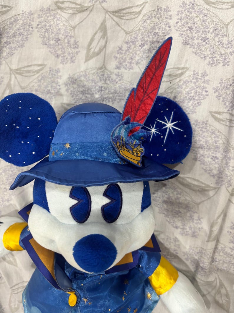 May include: A plush Mickey Mouse dressed as a sorcerer, wearing a blue hat with a red feather and a gold emblem. The ears are dark blue with white star accents. The outfit is blue and gold.