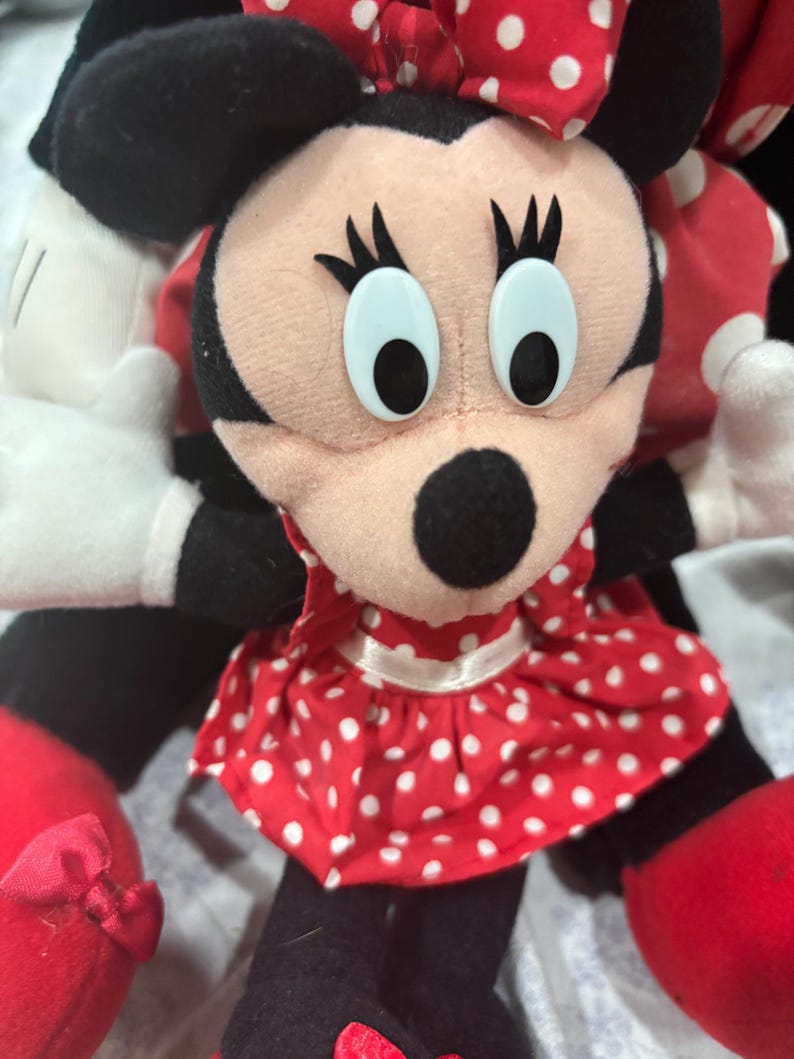 May include: A Minnie Mouse plush doll with a red and white polka dot dress and bow. The doll has black ears, a black nose, and large, light blue eyes. The doll's arms and legs are black, and it has red shoes.