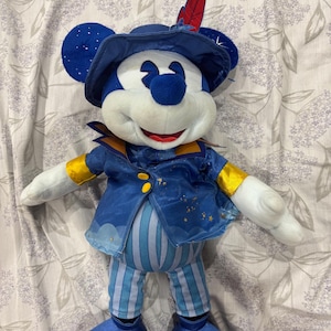 May include: A plush Mickey Mouse doll wearing a blue outfit with gold accents. The doll has a hat with a red feather and a jacket with gold buttons. The pants are blue and white striped. The doll's face is white with a red smile and a blue nose.