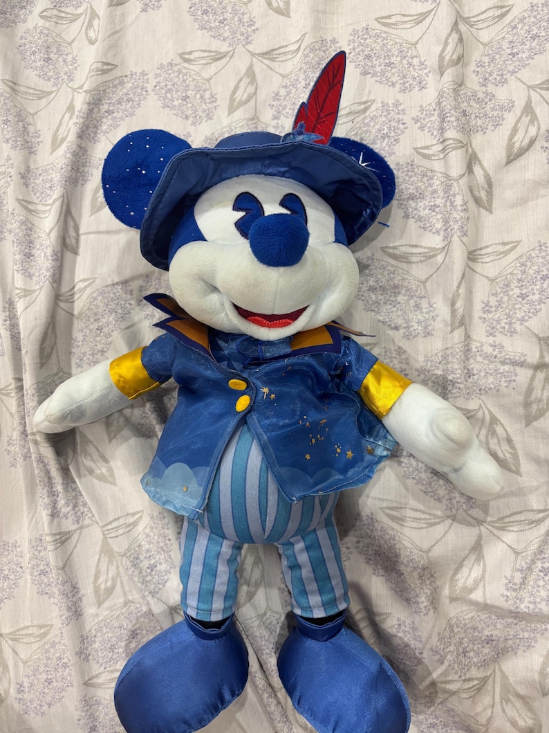 May include: A plush Mickey Mouse doll wearing a blue outfit with gold accents. The doll has a hat with a red feather and a jacket with gold buttons. The pants are blue and white striped. The doll's face is white with a red smile and a blue nose.