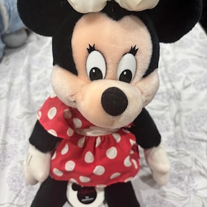May include: A plush Minnie Mouse doll featuring a black head and ears, a cream-colored bow, and a red dress with white polka dots. The doll has black legs, red shoes, and white gloves. The doll is sitting on a white surface.