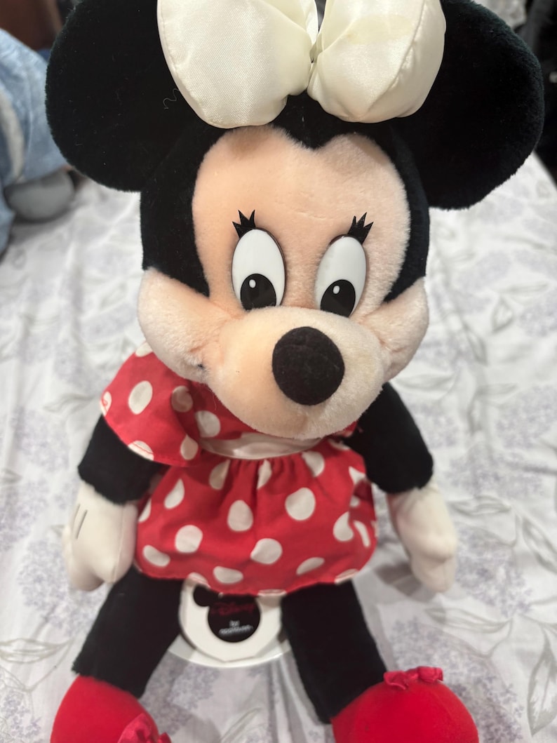 May include: A plush Minnie Mouse doll featuring a black head and ears, a cream-colored bow, and a red dress with white polka dots. The doll has black legs, red shoes, and white gloves. The doll is sitting on a white surface.
