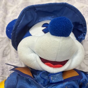 May include: A plush toy of a character wearing a blue hat and outfit. The character has a white face, blue ears, and a red mouth. The outfit has gold accents and yellow sleeves. The toy is soft and cuddly.