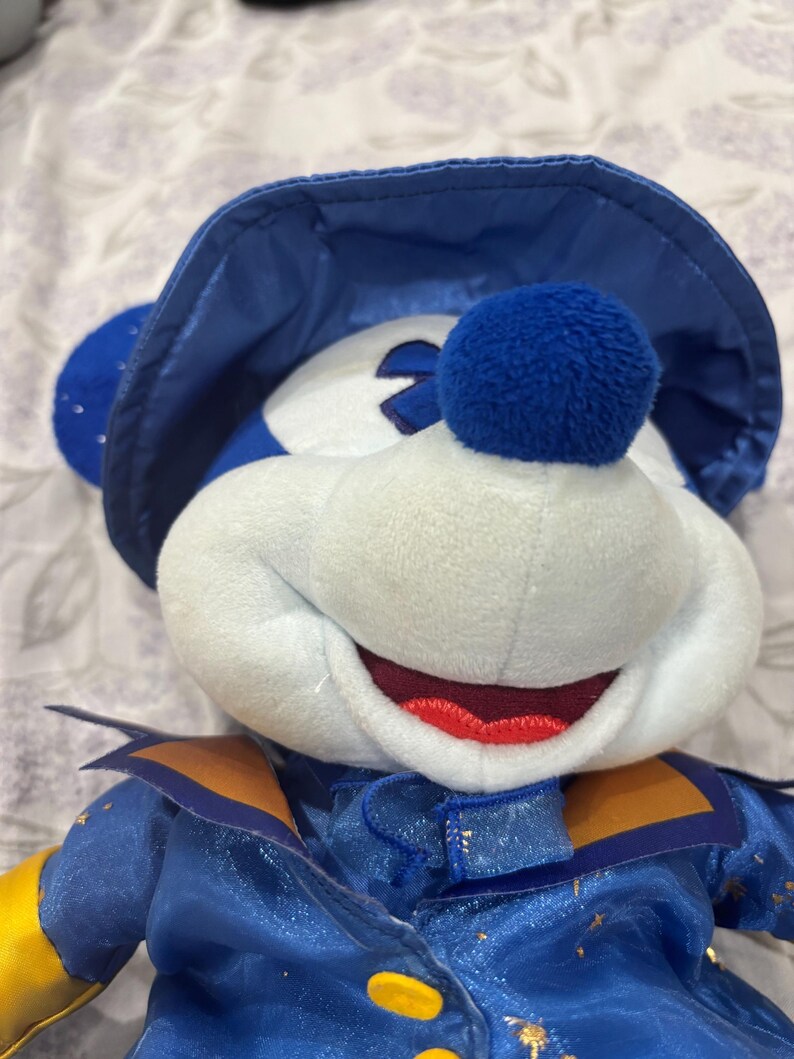 May include: A plush toy of a character wearing a blue hat and outfit. The character has a white face, blue ears, and a red mouth. The outfit has gold accents and yellow sleeves. The toy is soft and cuddly.