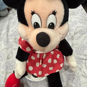 May include: A plush Minnie Mouse doll with a black head and ears, a pink face, and large black eyes. She wears a red dress with white polka dots and a white bow. Her arms and legs are black and white, with red shoes.