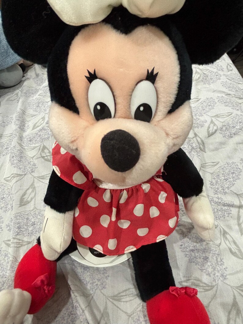 May include: A plush Minnie Mouse doll with a black head and ears, a pink face, and large black eyes. She wears a red dress with white polka dots and a white bow. Her arms and legs are black and white, with red shoes.