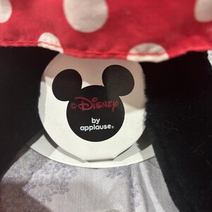 May include: A close-up of a Minnie Mouse plush toy. The toy features a red polka-dot bow and black ears. The Disney by Applause tag is visible. The plush is made of soft materials.