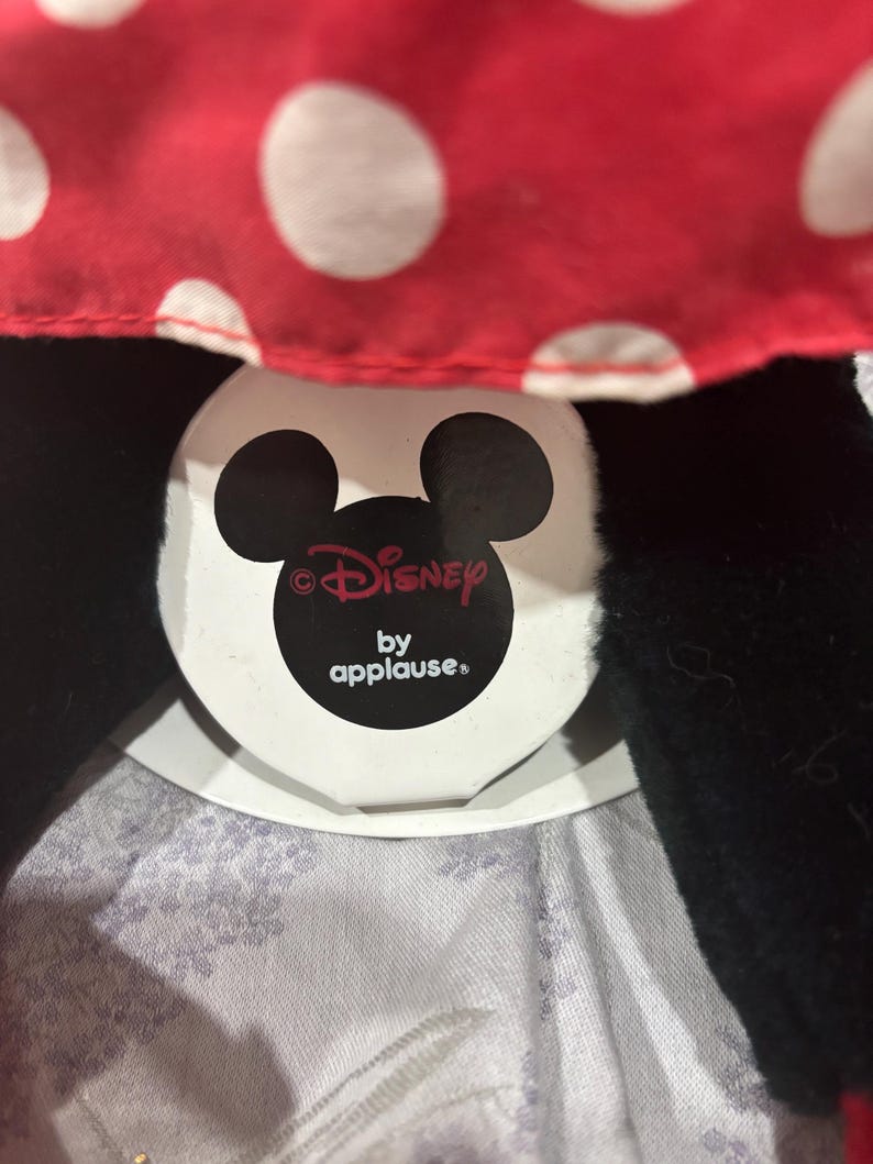 May include: A close-up of a Minnie Mouse plush toy. The toy features a red polka-dot bow and black ears. The Disney by Applause tag is visible. The plush is made of soft materials.