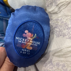 May include: Close-up of a blue plush toy foot with a circular patch. The patch features the text "Mickey Mouse The Main Attraction Series 6 of 12" with a colorful Mickey Mouse graphic. The plush toy is light blue and white.