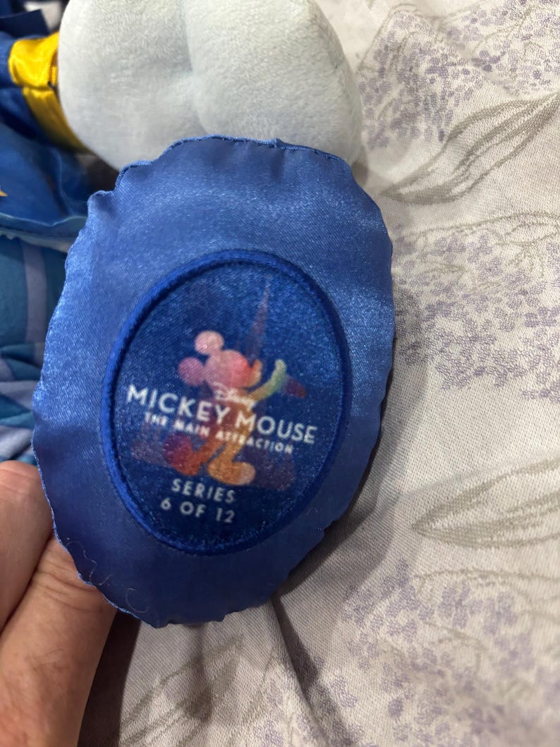 May include: Close-up of a blue plush toy foot with a circular patch. The patch features the text "Mickey Mouse The Main Attraction Series 6 of 12" with a colorful Mickey Mouse graphic. The plush toy is light blue and white.