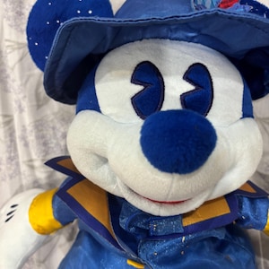 May include: A plush Mickey Mouse doll wearing a blue hat with a feather, a blue and gold jacket, and white gloves. The doll has a blue nose and eyes, and white face. The hat has silver sparkles.