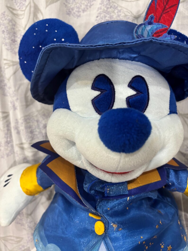 May include: A plush Mickey Mouse doll wearing a blue hat with a feather, a blue and gold jacket, and white gloves. The doll has a blue nose and eyes, and white face. The hat has silver sparkles.