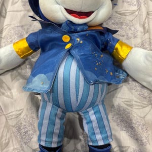 May include: A plush toy of a mouse character wearing a blue outfit with gold accents. The outfit includes a jacket with star details, striped pants, and a hat. The mouse has white fur, a red mouth, and large blue feet.