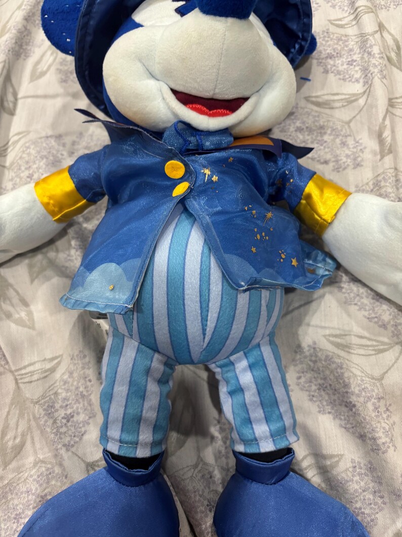 May include: A plush toy of a mouse character wearing a blue outfit with gold accents. The outfit includes a jacket with star details, striped pants, and a hat. The mouse has white fur, a red mouth, and large blue feet.