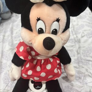 May include: A plush Minnie Mouse doll with black ears, a white bow, and a red dress with white polka dots. The doll has black legs, white gloves, and red shoes. The doll is sitting on a white surface.