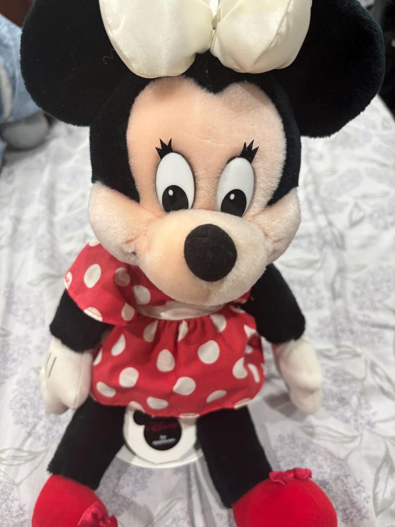 May include: A plush Minnie Mouse doll with black ears, a white bow, and a red dress with white polka dots. The doll has black legs, white gloves, and red shoes. The doll is sitting on a white surface.