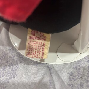 May include: A black plush toy with a red bow and white polka dots. The toy has a tag that reads "applause" and "Made in India". The toy is sitting on a white stand.