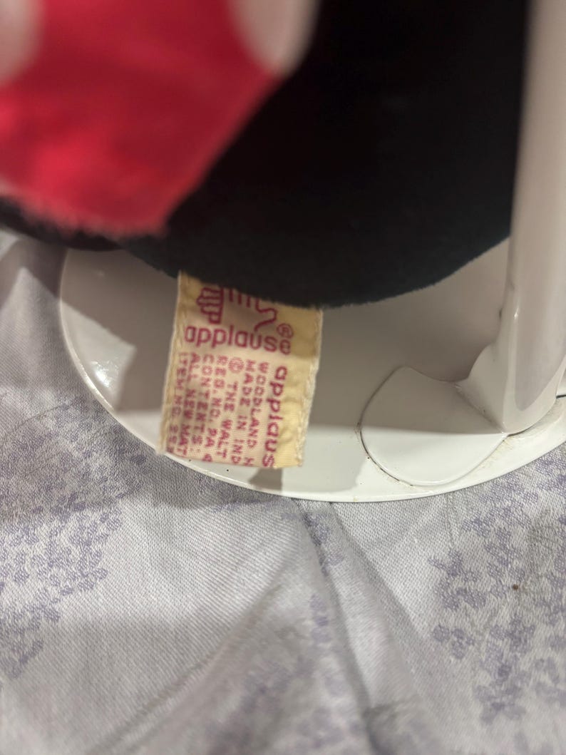 May include: A black plush toy with a red bow and white polka dots. The toy has a tag that reads "applause" and "Made in India". The toy is sitting on a white stand.