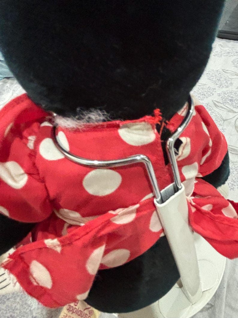 May include: A plush toy featuring a black head and a red dress with white polka dots. The dress is held by a metal hanger with a white plastic piece. The toy is a classic character.