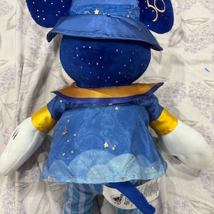 May include: A plush Mickey Mouse doll in a blue and gold outfit. The doll has a hat with gold stars and the text "Walt Disney World 50". The doll's outfit includes a blue tunic with gold accents and striped pants.