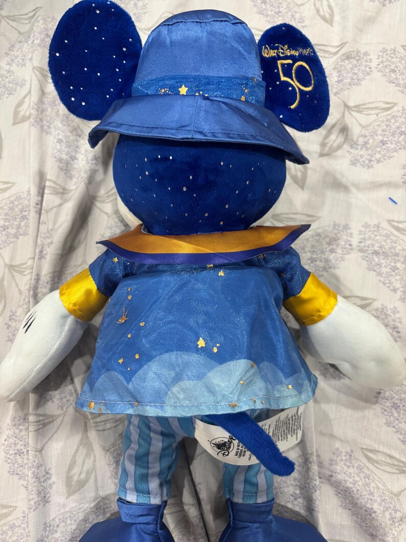 May include: A plush Mickey Mouse doll in a blue and gold outfit. The doll has a hat with gold stars and the text "Walt Disney World 50". The doll's outfit includes a blue tunic with gold accents and striped pants.