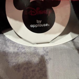 May include: A tag featuring the Disney logo and the text "by applause." The tag is black with a white border and a Mickey Mouse head shape. The top of the image shows a red and white striped fabric.