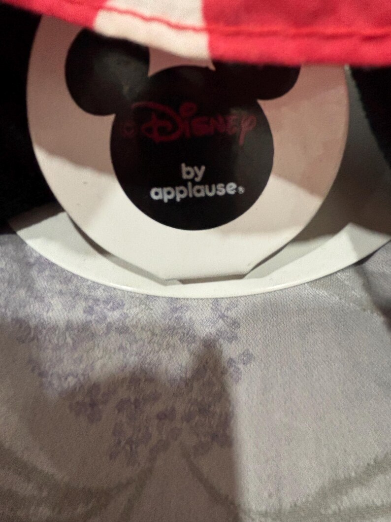 May include: A tag featuring the Disney logo and the text "by applause." The tag is black with a white border and a Mickey Mouse head shape. The top of the image shows a red and white striped fabric.
