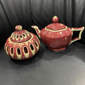 May include: A red ceramic teapot and a matching round container with a lid. The teapot has a curved spout and handle with a light green trim. The round container has oval cutouts and a matching lid. Both items are on a black surface.