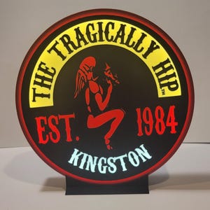 May include: A round, black and red sign with the words "The Tragically Hip" in yellow lettering. The sign also features a red silhouette of a figure with wings, the words "Est. 1984", and "Kingston" in white lettering.
